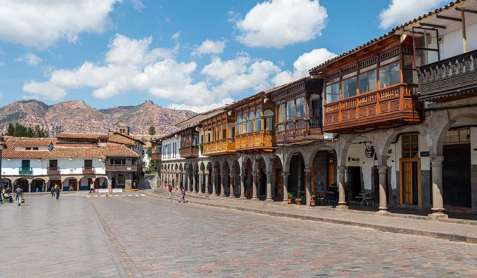 Historische Kolonialgebäude mit Holzbalkonen und Steinarkaden am Plaza de Armas in Cusco, Peru, bei sonnigem Wetter mit Bergen im Hintergrund.