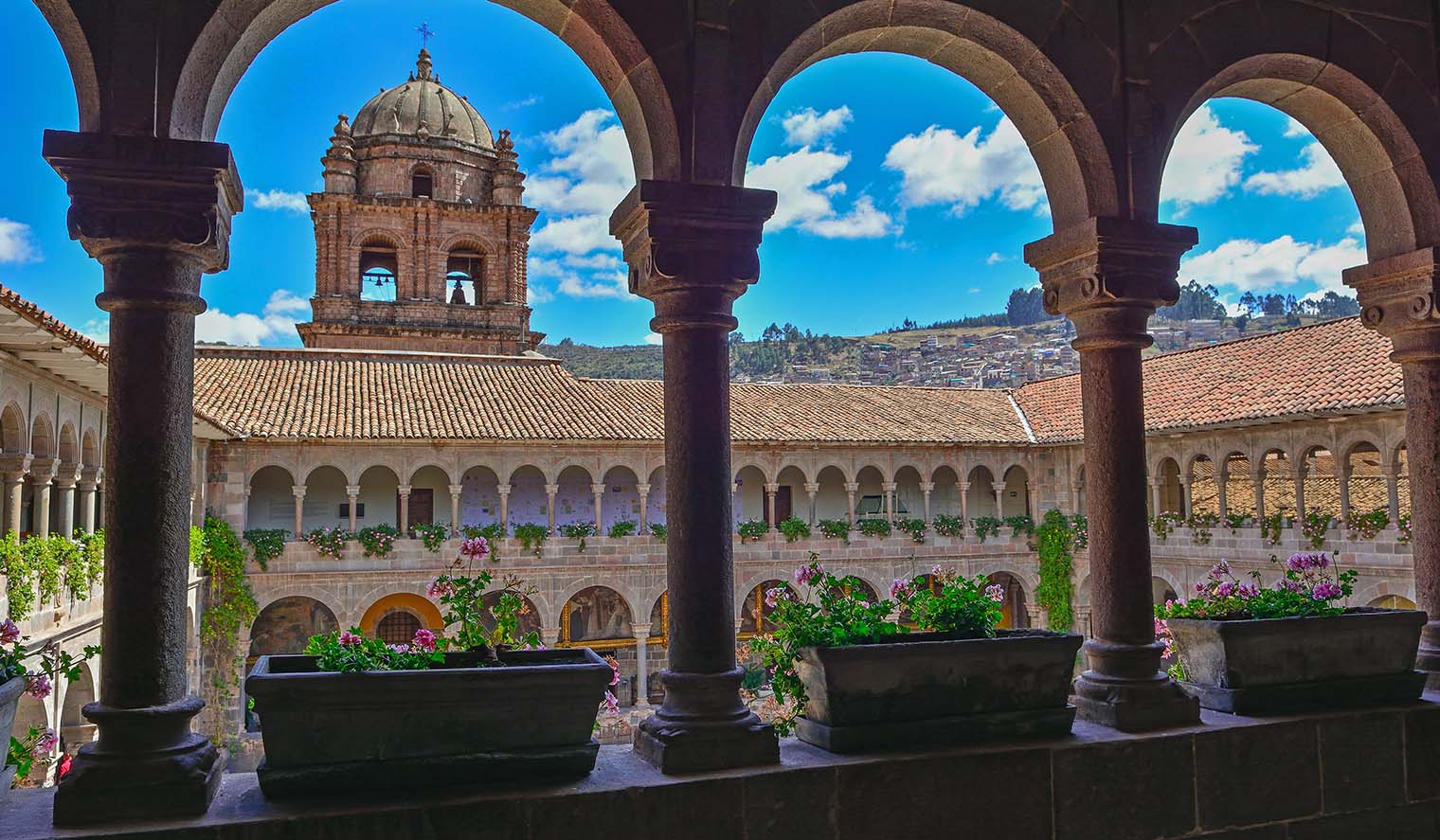 Blick durch steinerne Rundbögen auf den blumengeschmückten Innenhof des Klosters Santo Domingo in Cusco, Peru, mit der barocken Kirchturmkuppel und Hügeln im Hintergrund.